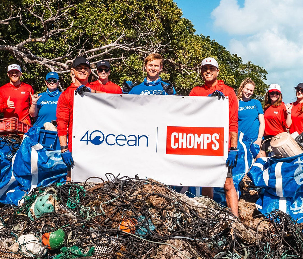 A group of volunteers wearing red and blue 4Ocean CHOMPS uniforms stand behind a large white banner with the 4Ocean and CHOMPS logos, holding a massive pile of fishing nets and debris outdoors under a blue sky with trees in the background.