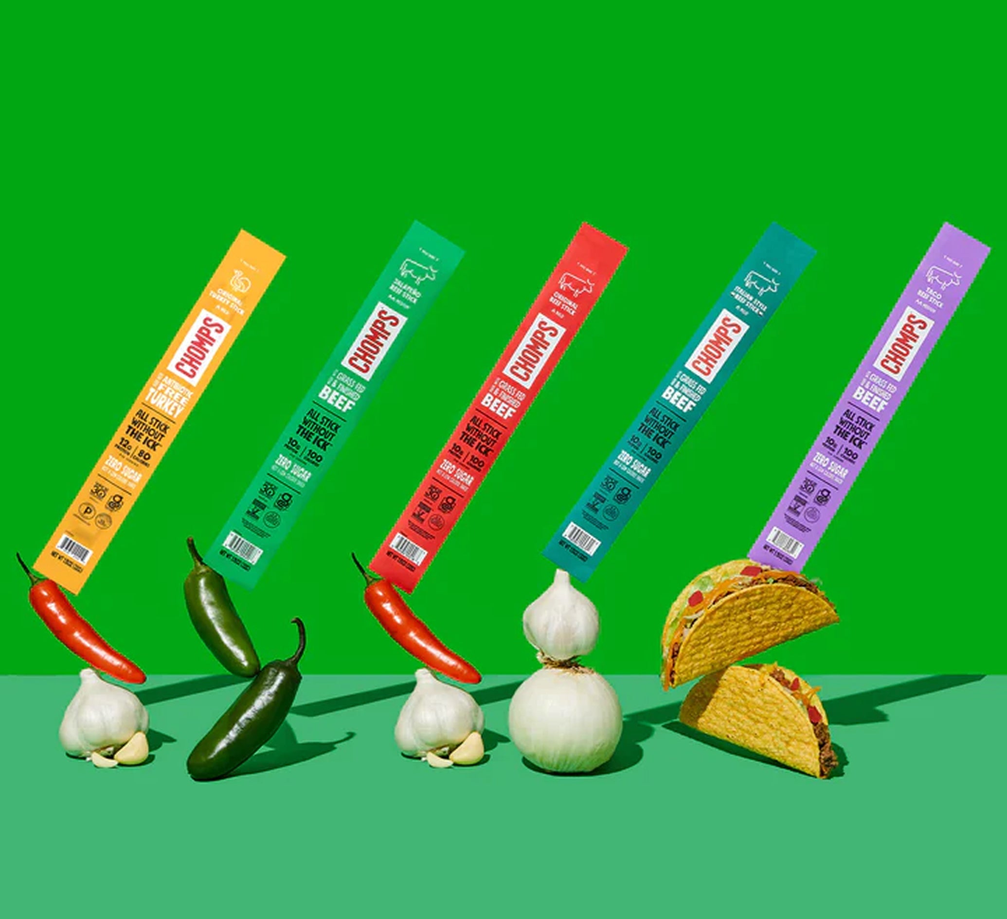 Four colorful Chomps sticks are arranged diagonally on a bright green background, with corresponding food props below: red and green chili peppers, garlic bulbs, and two tacos.