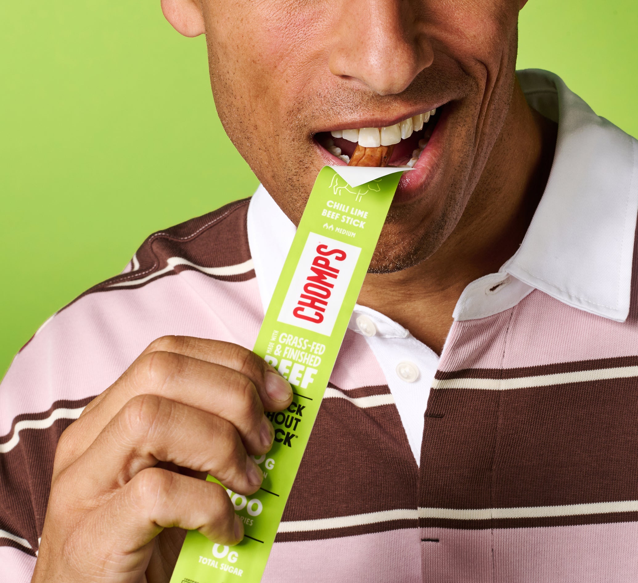 A close-up lifestyle shot of a man biting into a Chomps Chili Lime beef stick against a solid green background