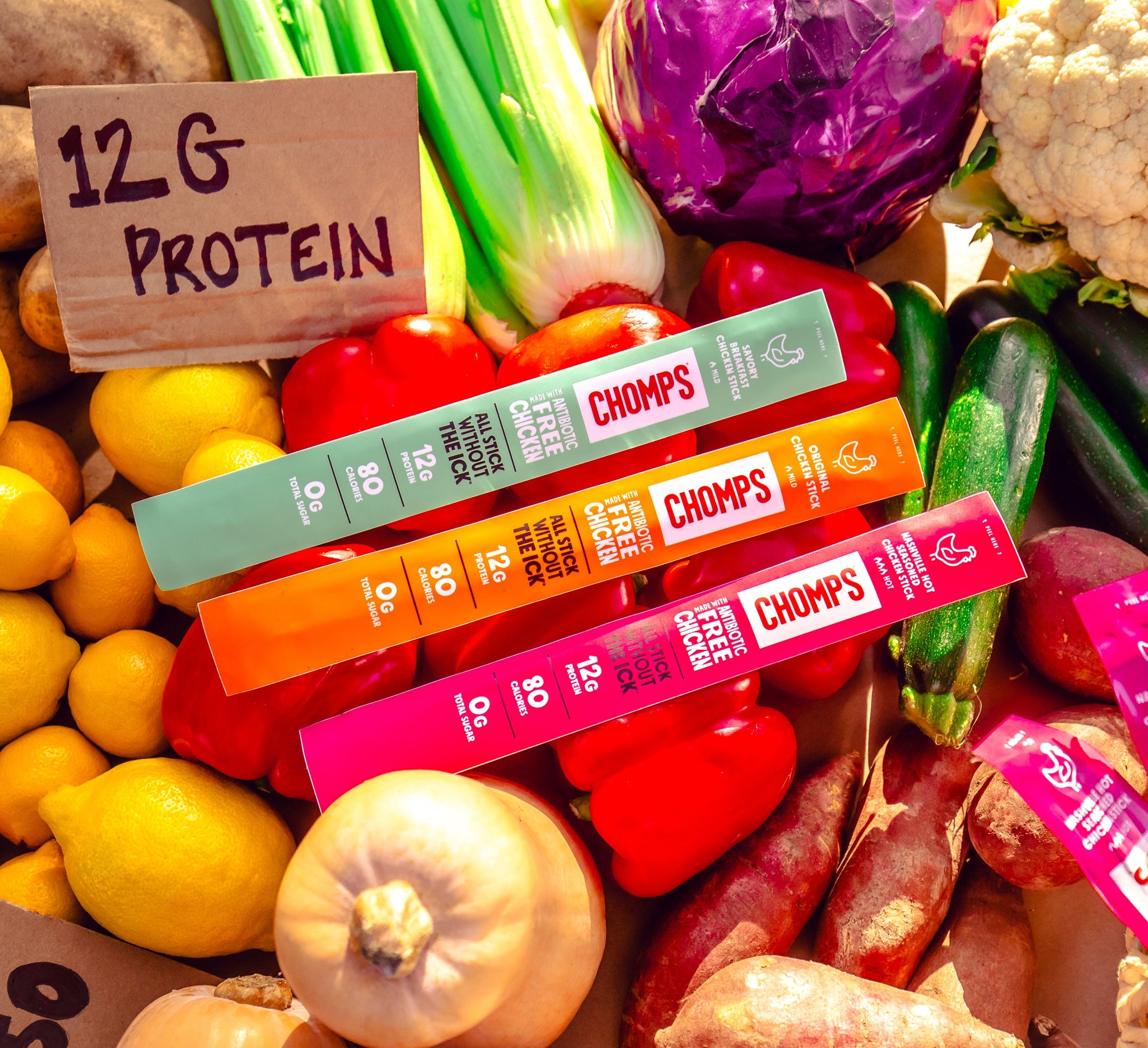 Three Chomps sticks arranged on a colorful background of fresh vegetables and fruits, including lemons, onions, peppers, and cabbage. A handwritten paper sign reads '12G PROTEIN'.