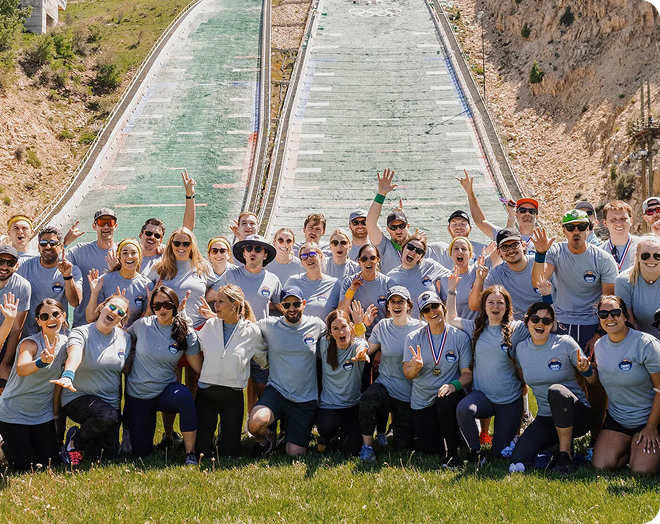 Chomps employees posing together outdoors in front of a long ski slope, wearing matching light blue t-shirts with the Chomps logo