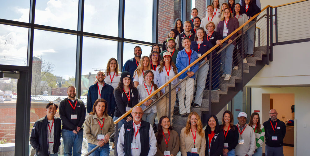 A group of people posing together on an indoor staircase in a modern building with large windows. Most are wearing matching red lanyards and badges.