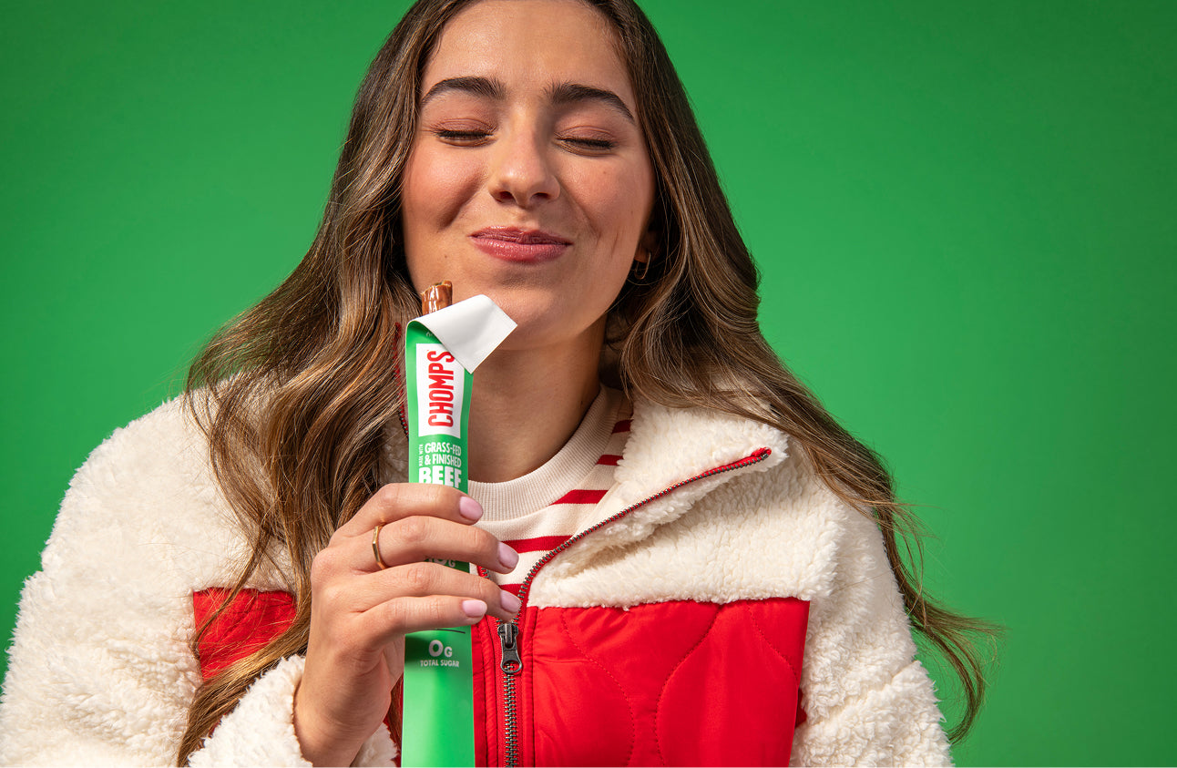 A woman with long brown hair holds a Chomps Jalapeno Beef stick against a solid green background. She is wearing a white sherpa-lined puffer jacket with red panels.