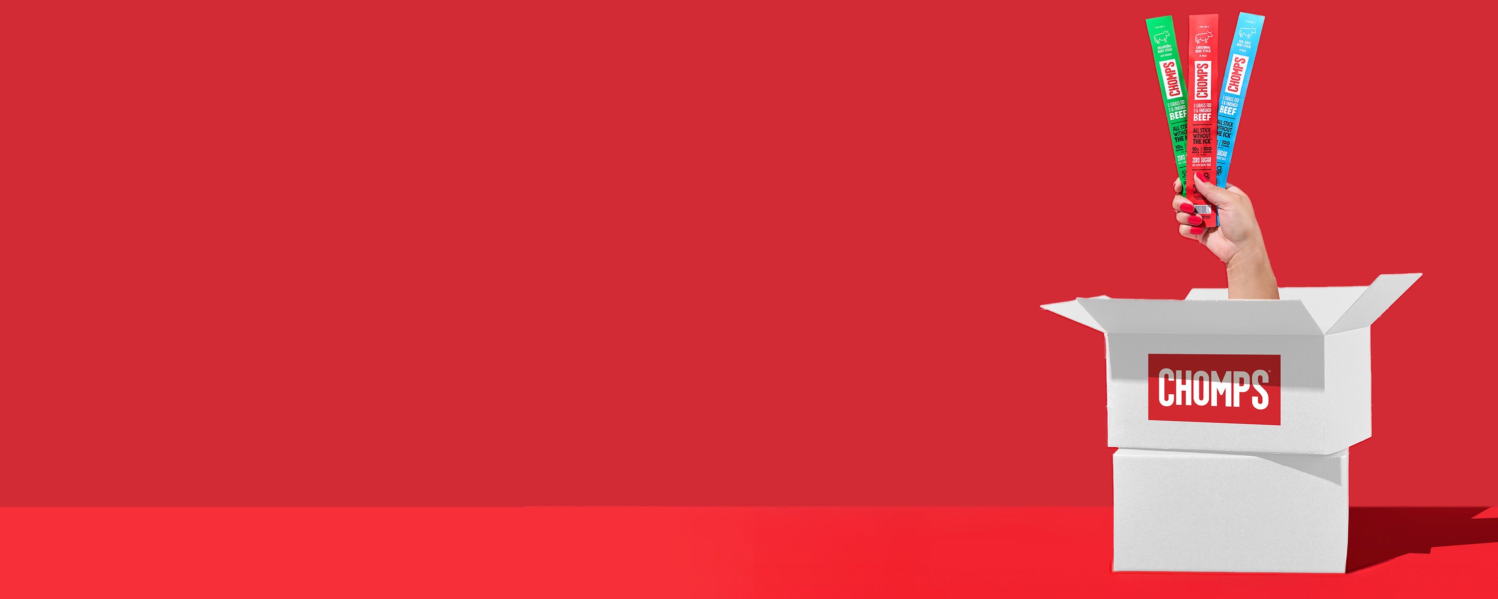 Hand holding an assortment of Chomps above a white box with 'CHOMPS' branding on a red background