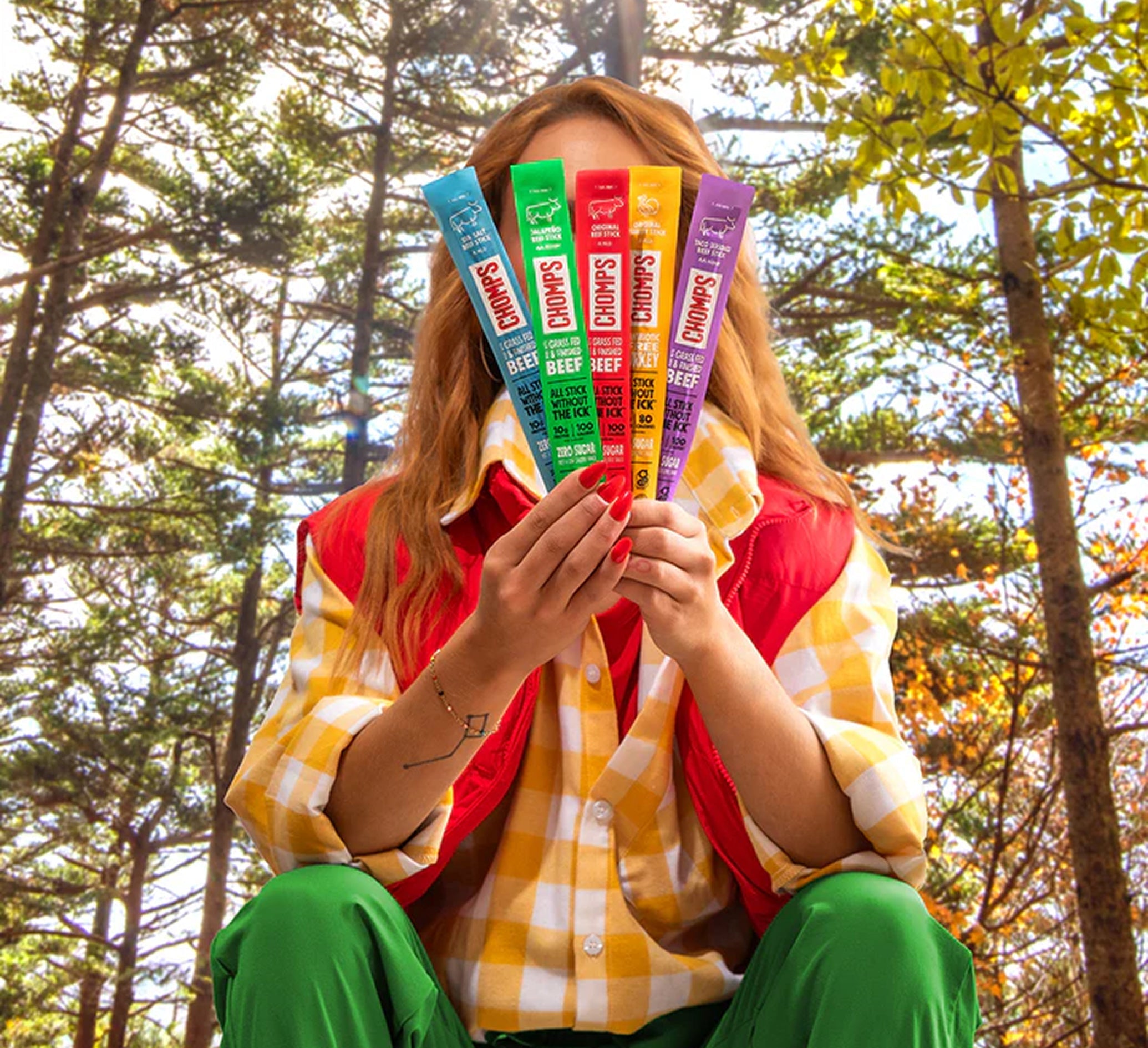 A person sitting outdoors in a wooded area holds five colorful Chomps sticks The person is wearing a yellow and white checkered shirt, a red vest, and green pants.