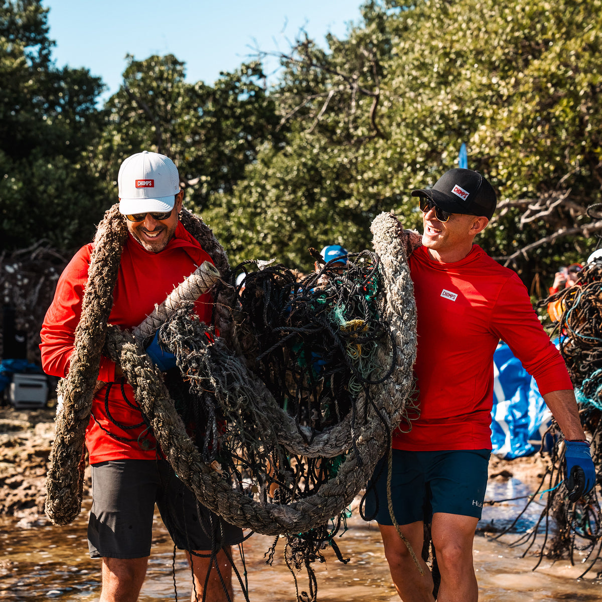 Pete and Rashid in red athletic shirts and shorts carry a large tangled fishing net through shallow water outdoors, with trees and blue sky in the background.