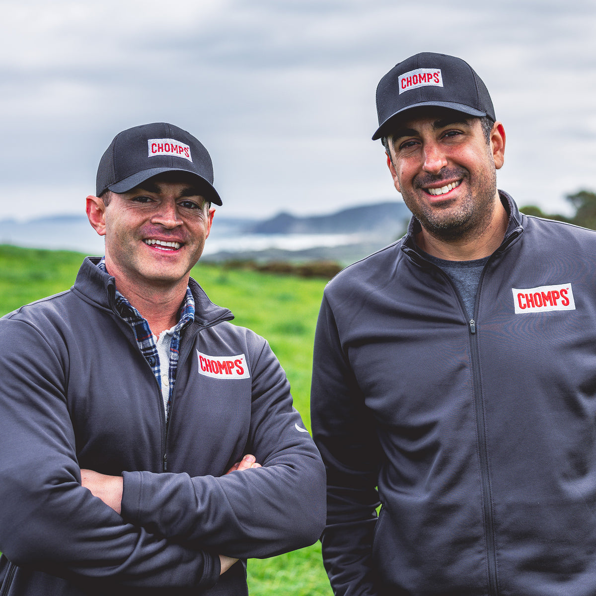Pete and Rashid standing outdoors in a grassy field under an overcast sky, wearing matching dark gray CHOMPS branded zip-up jackets and baseball caps.