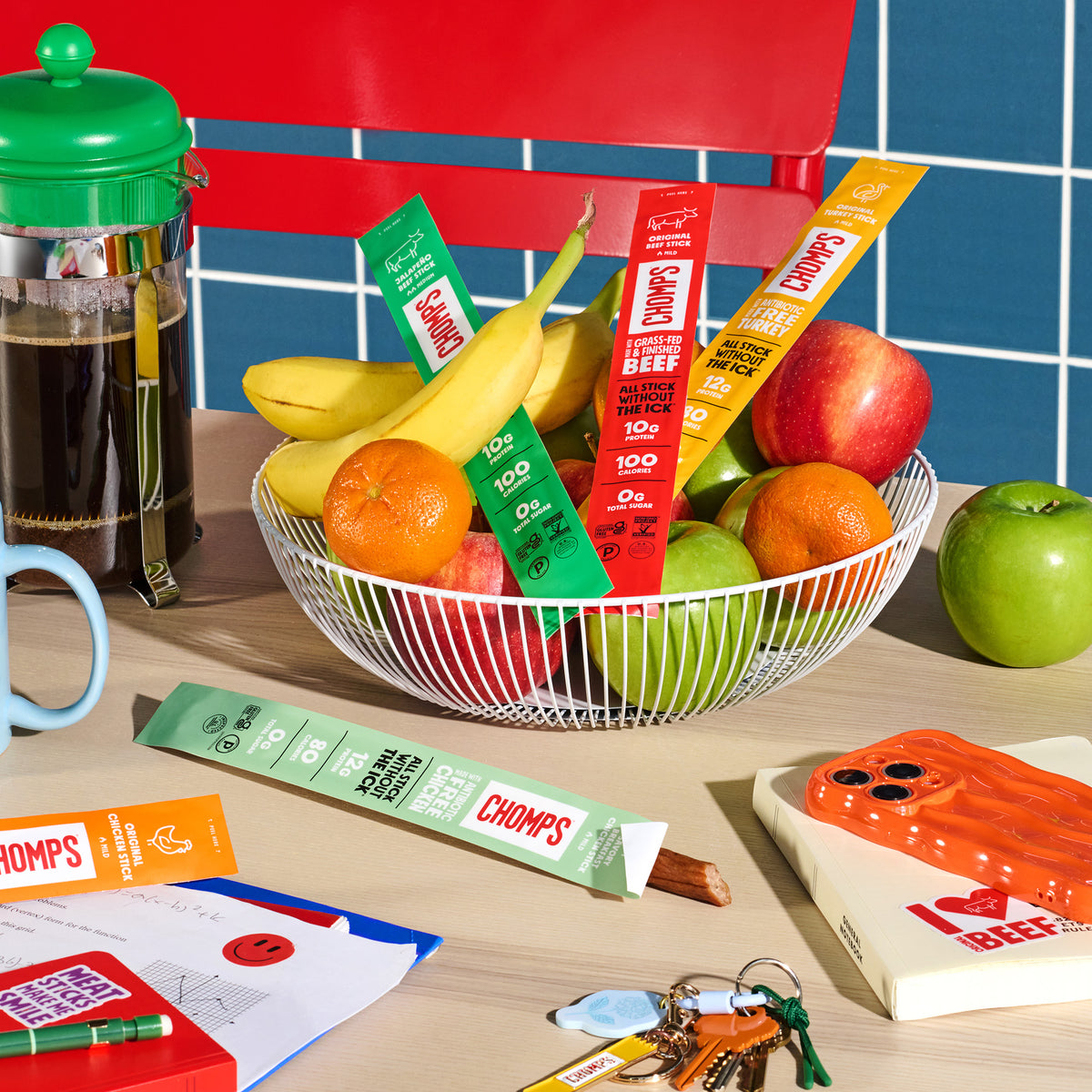 A styled tabletop scene with a wire bowl filled with fruit and several colorful snack sticks labeled CHOMPS. A coffee maker, mug, keys, notebook, and other breakfast props are arranged around it against a blue tiled wall.