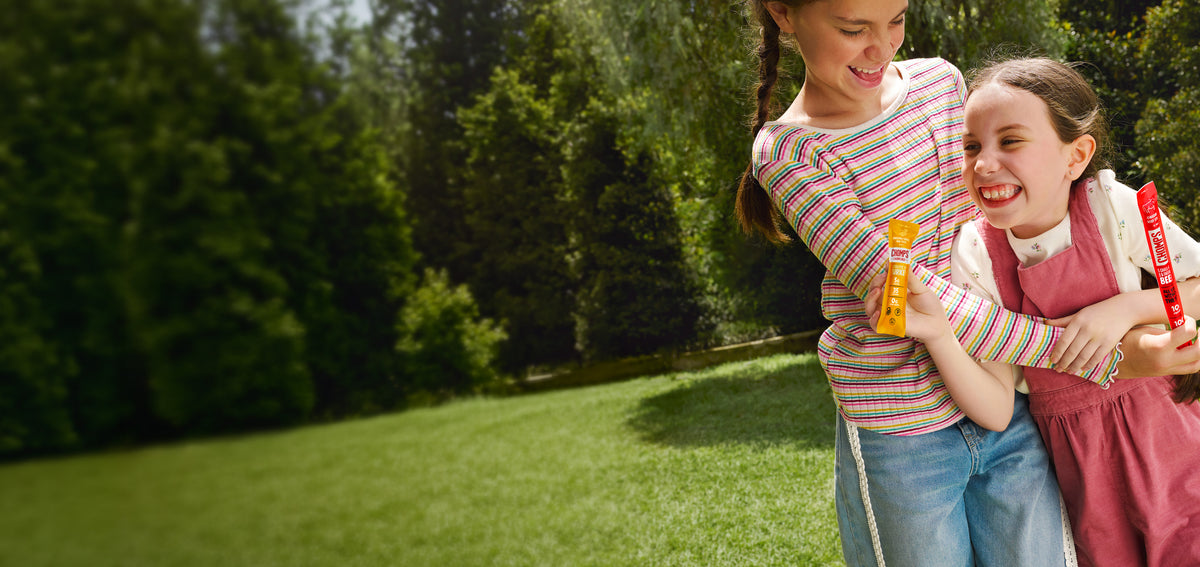 Two young girls hugging outdoors on a grassy lawn with trees in the background. The girl on the left wears a pink, yellow, and blue striped long-sleeve shirt and blue jeans and is holding a Chomps Beef stick. The girl on the right is wearing a pink dress and is holding a Chomps turkey stick product.