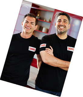 Pete and Rashid wearing matching black short-sleeve t-shirts with a red and white 'Chomps' logo on the chest, standing in a kitchen-like setting with red shelving in the background.