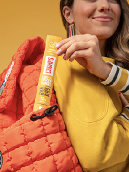 A woman in a yellow sweatshirt and striped sleeve holds a Chomps sick tucked into an orange quilted puffer tote against a warm yellow background.
