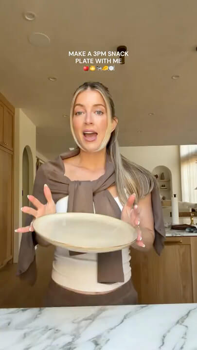 A woman in a kitchen setting holds up a round beige plate, with text overlay that reads 'MAKE A 3PM SNACK PLATE WITH ME'.
