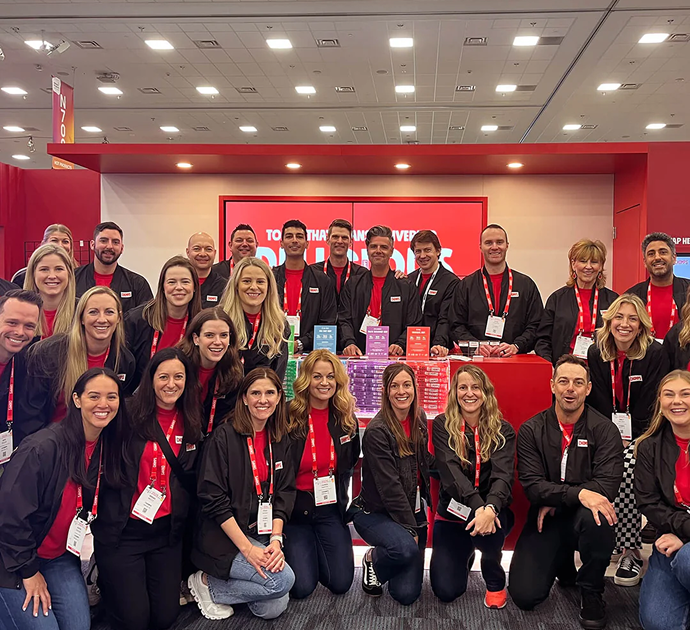 A large group of people posing together wearing matching Chomps black jackets and red shirts, with lanyards and colorful Chomps product boxes in front of a red display wall.