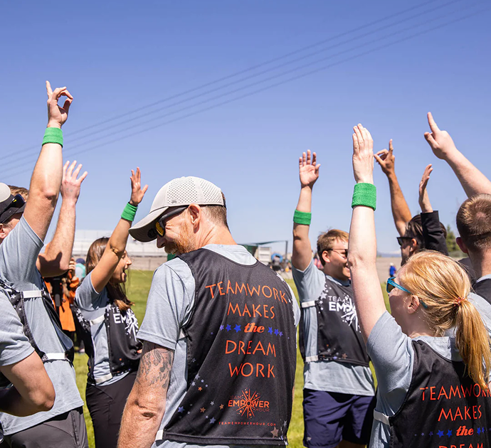 A group of people outdoors wearing black vests printed with the text 'TEAMWORK MAKES the DREAM WORK' and 'EMPOWER' in orange and white, with several people raising their hands under a blue sky.