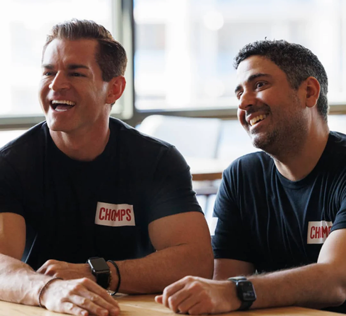 Pete and Rashid smiling and laughing at a table, both wearing matching black t-shirts with a white square logo on the left chest that reads 'Chomps' in red letters.