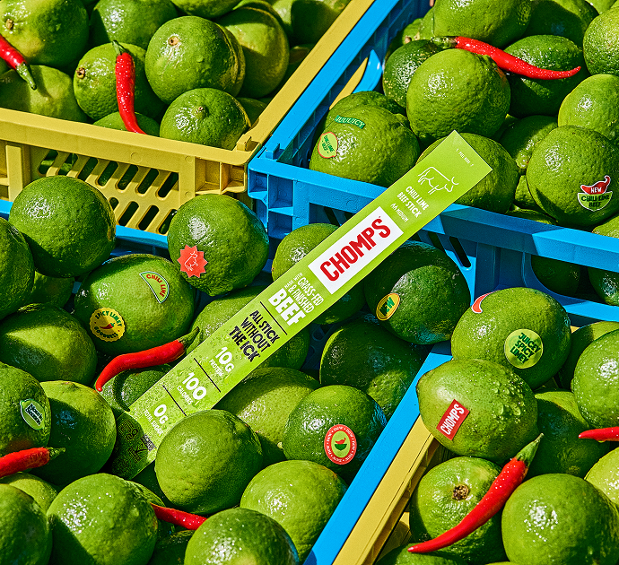 A close-up overhead view of crates filled with bright green limes, with a Chomps Chili Lime Beef stick placed across the crates and a few red chili peppers scattered among the limes.