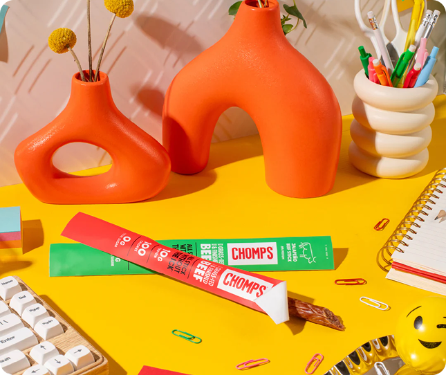 A styled desk scene featuring a package of Chomps snack sticks, with two orange sculptural vases, a cream pen cup, and other desk accessories on a bright yellow surface.