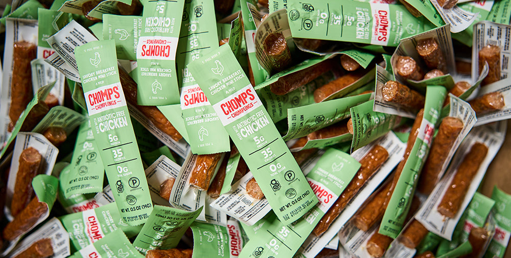 A close-up pile of Chomp's Savory Breakfast chicken jerky sticks in their light green and white individual packaging, with the brown jerky visible through clear plastic wrapping.