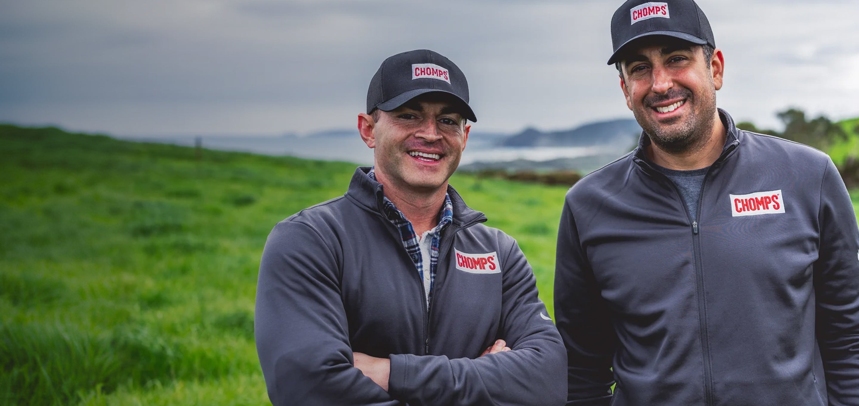Pete and Rashid standing outdoors in a grassy field under an overcast sky, both wearing dark gray zip-up jackets and matching dark baseball caps with a red and white CHOMP'S logo on the chest and cap.