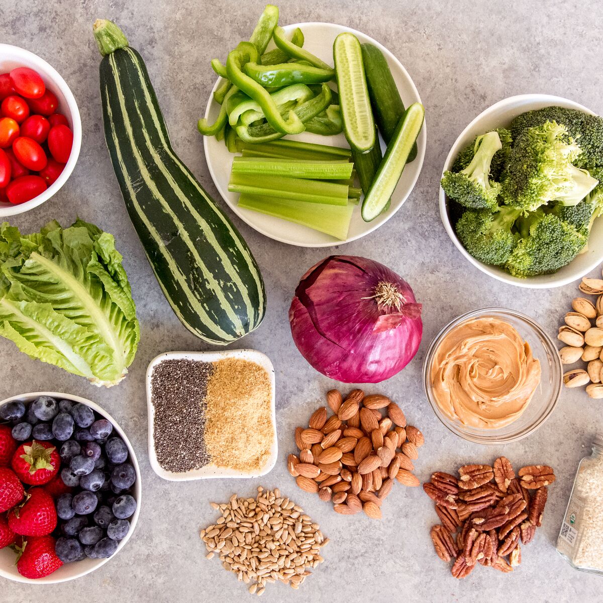 An assortment of keto friendly ingredients: cucumbers, peppers, broccoli, cherry tomatoes, zucchini, spices, blueberries, strawberries, and nuts