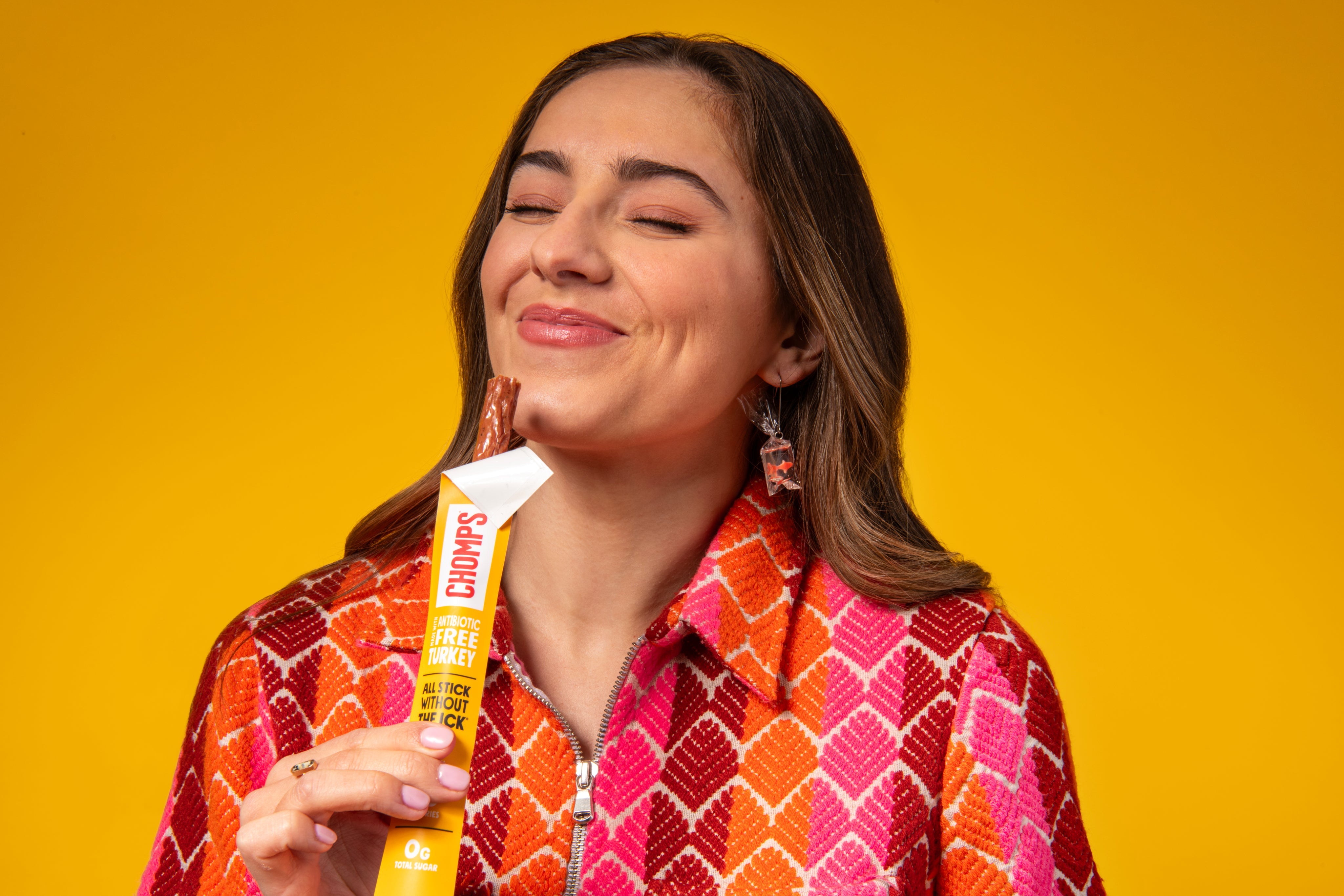 A woman with her eyes closed holds an Original Turkey meat stick, showing the product partially pulled from its wrapper. She is wearing a vibrant orange, pink, and red patterned shirt against a solid yellow background.